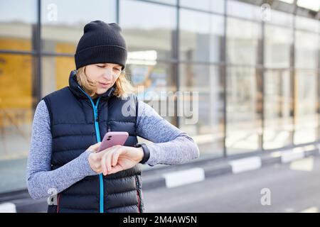 Frau mit Mobiltelefon, das an eine Smart Watch angeschlossen ist. Die Sportlerin schaut auf die Smartwatch und hält ihr Smartphone in der anderen Hand, im Freien. Fitness-FEM Stockfoto