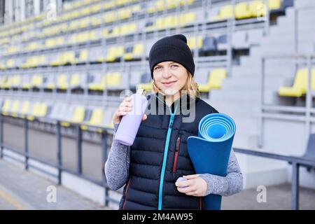 Junge attraktive lächelnde Frau, die morgens bei Sonnenaufgang im Park Sport treiben, Yogamatte und eine Flasche Wasser in der Hand halten, gesunder Lebensstil Stockfoto