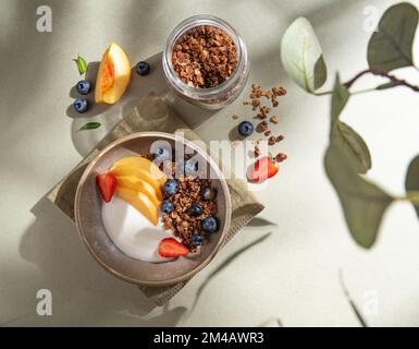 Köstlicher natürlicher Joghurt mit hausgemachtem Müsli, Pfirsich, Beeren in einer Schüssel und einem Glas mit Müsli auf grünem Hintergrund im Morgenschatten. Gesund und verrückt Stockfoto