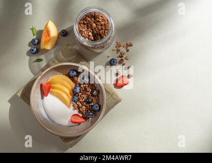 Köstlicher Naturjoghurt mit hausgemachtem Müsli, Pfirsich, Beeren in der Schüssel und einem Glas mit Müsli auf grünem Hintergrund im Morgenschatten. Gesund und nahrhaft Stockfoto