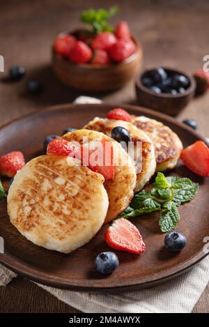 Hüttenkäse-Pfannkuchen oder Syrniki mit Minze, Erdbeeren und Blaubeeren auf einem dunklen Holztisch. Konzept des gesunden Frühstücks. Makro und selektiv Stockfoto