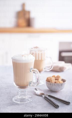 Zwei Gläser duftender Cappuccino-Kaffee mit Zimt und braunem Zucker auf einem blauen Tisch vor dem Hintergrund einer weißen Küche am frühen Morgen Stockfoto