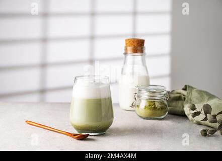 Grüner Matcha Latte in einem Glas auf grauem Hintergrund mit Morgenschatten aus dem Fenster, eine Flasche Milch und Teepulver das Konzept eines veganen Getränks, Stockfoto