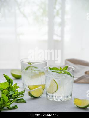 Zwei Gläser Limettenmojito auf dem Tisch mit Limettenscheiben und Minze im Morgenlicht vom Fenster. Vorderansicht Stockfoto