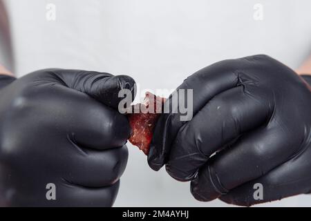 Ein Typ in schwarzen Handschuhen zerbricht ein Stück getrocknetes, gepökeltes Fleisch auf weißem Hintergrund. Hochwertiges Foto Stockfoto