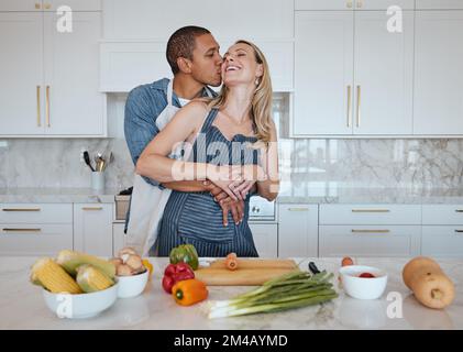 Ein Paar kocht Essen und liebt Gemüse mit einem Kuss auf die Wange, während er beim Abendessen zu Hause hilft. Mann und Frau in Großbritannien mit einem gesunden Lebensstil Stockfoto