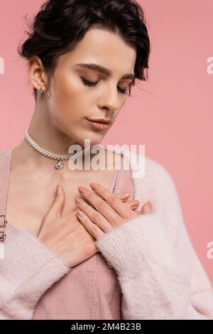 Junge Frau in Perlenkette und Piercing Pose mit Händen auf der Brust und geschlossenen Augen isoliert auf rosa Stockfoto