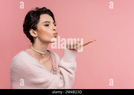 Trendige Frau in flauschiger Strickjacke und Perlenkette, die einen Luftkuss versendet, während sie isoliert in Pink wegschaut Stockfoto