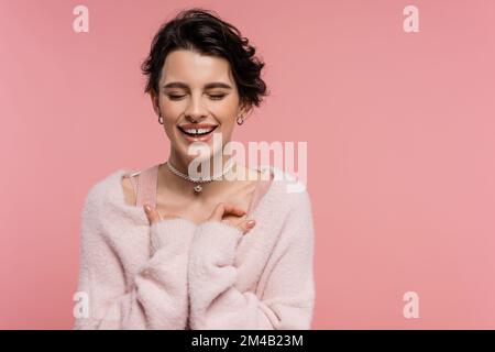 Fröhliche Frau in flauschiger Strickjacke und Perlenkette, die lacht, mit geschlossenen Augen, isoliert auf Pink Stockfoto