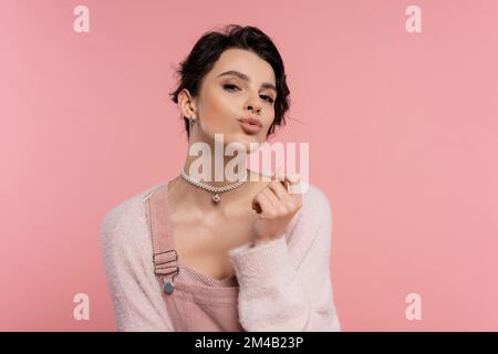 Stilvolle brünette Frau in Perlenkette, die Lippen schmollt und auf die Kamera schaut, isoliert auf Pink Stockfoto