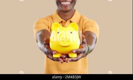 Schweinebank in Form eines gelben Schweines in den Händen eines lächelnden schwarzen Mannes auf beigem Hintergrund. Stockfoto