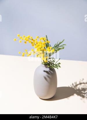 Ein Strauß gelber Mimosablüten steht in einer Keramikvase mit Schatten auf einem gelb-grauen Hintergrund. Konzept vom 8. März, fröhlicher Frauentag. Verstanden Stockfoto