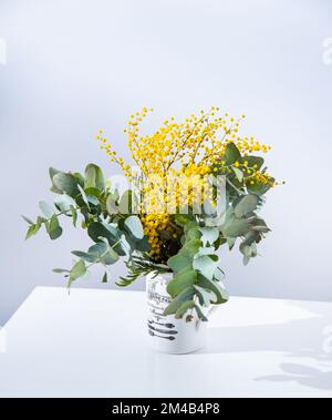 Ein Frühlingsstrauß mit gelben Mimosablüten und Eukalyptusästen steht auf einem weißen Tisch. Konzept vom 8. März, fröhlicher Frauentag. Speicherplatz kopieren Stockfoto