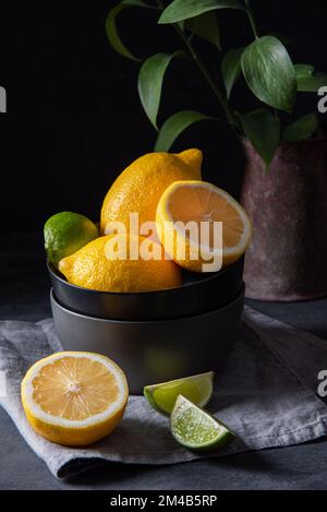Saftige Zitronen und Limetten in einer schwarzen Schüssel auf einem grauen Tisch. Dunkles und stimmungsvolles Konzept. Vorderansicht Stockfoto