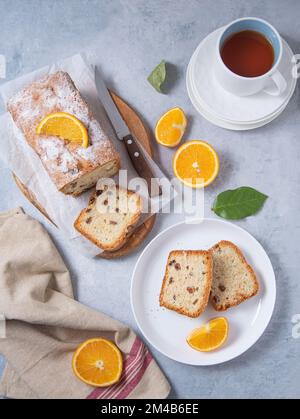 Konzept-Frühstück. Frisch gebackener Kuchen mit Rosinen und Orange auf einem Schneidebrett und blauem Hintergrund mit Obstorangen und einer Tasse Tee. Draufsicht i Stockfoto