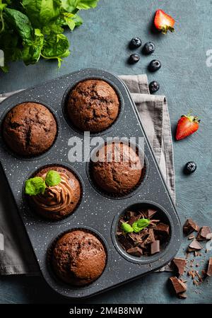 Schokoladen-Muffins mit Heidelbeeren- und Erdbeerscheiben in einem Backblech auf dunklem Hintergrund. Draufsicht. Stockfoto