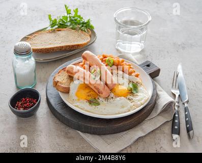 Traditionelles englisches Frühstück mit Spiegeleiern, Würstchen, Bohnen, frischen Kräutern, rotem Pfeffer und Toast auf grauem Hintergrund. Konzept für Morgenessen. Stockfoto