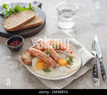 Traditionelles englisches Frühstück mit Spiegeleiern, Würstchen, Bohnen, frischen Kräutern, rotem Pfeffer und Toast auf grauem Hintergrund. Konzept für Morgenessen. Stockfoto