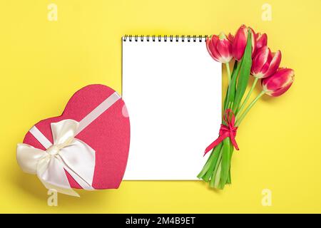 Strauß roter Tulpen, Geschenkbox Herz, Notizblock auf gelbem Hintergrund Top View Flat Lay Holiday Grußkarte Happy Moter's Day, 8. März, Valentinstag Stockfoto