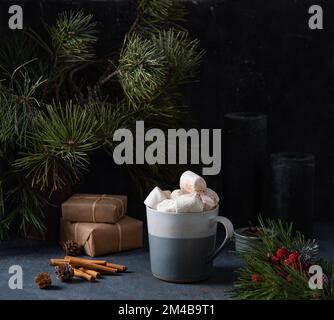 Tasse mit Kakao, Marshmallow auf dunkelblauem Hintergrund mit Zimt, Kerzen, Geschenken und Tanne. Dunkles und stimmungsvolles Bild Stockfoto