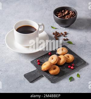 Köstliches Frühstück mit frisch gebrühtem Kaffee und Mini-Donuts in Puderzucker und Preiselbeeren auf schwarzem Schiefer auf blauem Hintergrund. Stockfoto