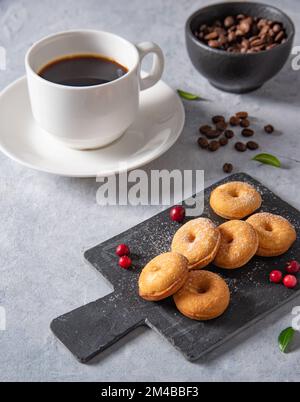 Köstliches Frühstück mit frisch gebrühtem Kaffee und Mini-Donuts in Puderzucker und Preiselbeeren auf schwarzem Schiefer auf blauem Hintergrund Stockfoto