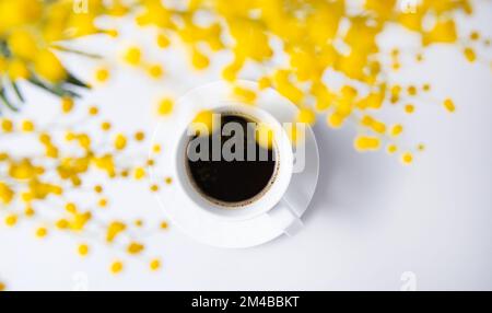 Ein Frühlingsstrauß mit gelben Mimosablüten über einer Tasse schwarzen Kaffees auf einem weißen Tisch. Konzept vom 8. März, fröhlicher Frauentag. Draufsicht und SPAC kopieren Stockfoto