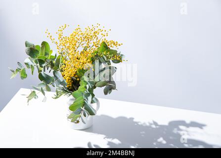 Ein Frühlingsbouquet mit gelben Mimosablüten und Eukalyptusästen steht auf einem weißen Tisch mit Schatten. Konzept vom 8. März, fröhlicher Frauentag. Kopie SPA Stockfoto