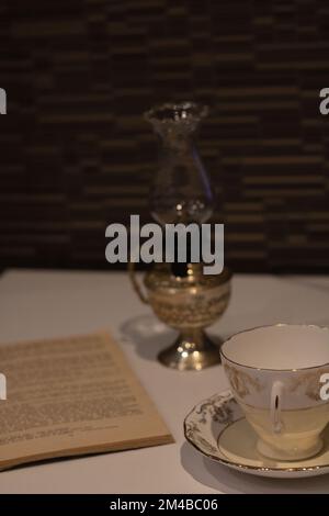 Foto eines offenen Buches und einer Kerosinlampe sowie einer leeren Tasse mit Untertasse, die auf dem Tisch steht Stockfoto