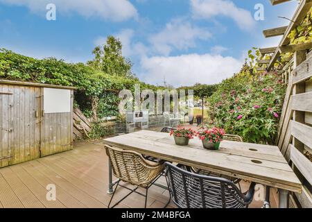 Eine Terrasse mit Holzmöbeln und Blumen auf dem Tisch im Hinterhof, umgeben von Büschen und Bäumen, gibt es ein Schild dafür Stockfoto