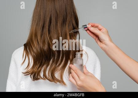 Haarstylisten, die Serum auf Haare junger Frauen auftragen, isoliert auf grau Stockfoto