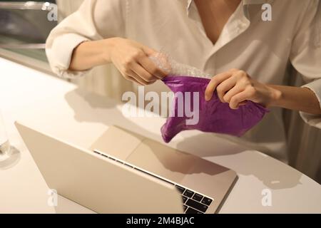 Frau öffnet Pakete, die in Luftpolsterfolie verpackt sind, während sie vor dem Laptop sitzt Stockfoto