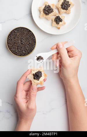 Schwarze Kaviar-Vorspeisen, Brotsterne mit Butter in menschlichen Händen auf weißem Hintergrund, Draufsicht Vertikal Stockfoto