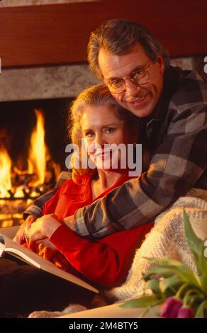 Ein älteres Paar, das sich umarmt und pausiert, um in die Kamera zu schauen, während es auf der Couch vor einem Kamin sitzt und zusammen ein Buch liest Stockfoto