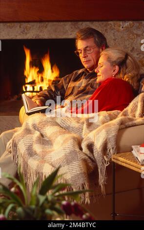 Ein älteres Paar, das auf der Couch saß, in eine Decke eingewickelt vor einem Kamin und ein Buch anschaute Stockfoto
