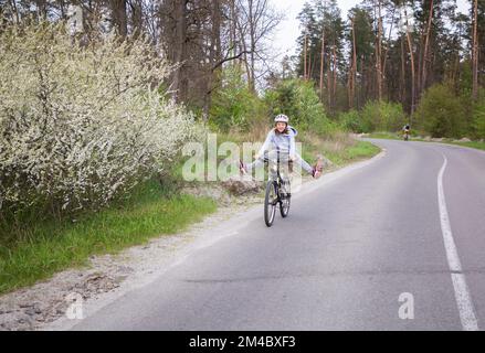 Glückliche Frau mit Helm fährt mit dem Fahrrad auf einer Asphaltstraße durch den Wald entlang blühender Bäume. Energie der Natur, Ruhe, Freiheit, Reisen. Aktiv Stockfoto
