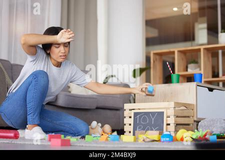 Spielzeug, Mutter müde und Kopfschmerzen beim Putzen, unordentliches Wohnzimmer und erschöpft. Frau, saubere Spielsachen und weiblicher Stress, frustriert und aufgebracht Stockfoto