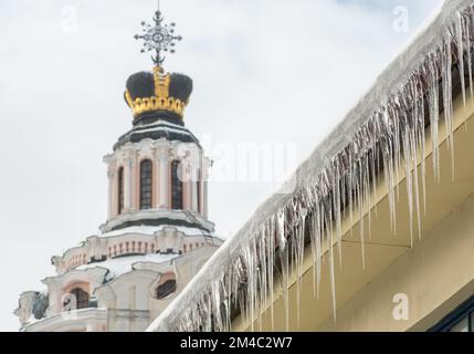 Eiszapfen und Schnee hängen an einem kalten Winter- oder Frühlingstag in Vilnius, Litauen, von einem Dach eines Gebäudes in der Stadt mit Kirche im Hintergrund Stockfoto