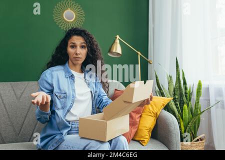 Porträt einer unzufriedenen Käuferin im Online-Shop, hispanische Frau, die enttäuscht von der Kamera war, die auf dem Sofa im Wohnzimmer saß, erhielt eine Schachtel Pakete per Post. Stockfoto