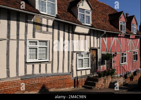 Erdbeere und Rahm … Die Fassaden dieser beiden Fachwerkhäuser wurden in den 1400s oder 1500s Jahren, als Lavenham in Suffolk, England, eine blühende Stadt für Wolle und Textilien war, wahrscheinlich als ein Grundstück erbaut. Grosvenor Cottage (Creme) und Old Rose Cottage (Erdbeerrosa) stehen auf Ziegelsteinplinden in der schrägen Prentice Street. Stockfoto