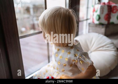 Mädchen mit 2 Jahren sitzt auf der Fensterbank. Eine kleine Blondine in einem weißen Kleid schaut aus dem Fenster. Stockfoto