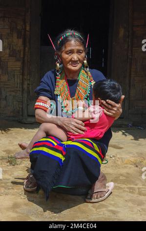 Mon, Nagaland, Indien - 03 03 2009 : Außenporträt der sitzenden alten Naga Konyak-Stammesfrau mit Enkelkind in traditionellem Kostüm und Schmuck Stockfoto