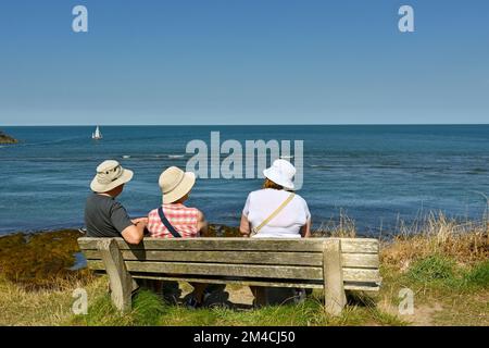 Newport, Pembrokeshire, Wales - 2022. August: Drei Personen sitzen auf einem Holzsitz und machen eine Pause von einem Spaziergang entlang des Küstenpfads Stockfoto