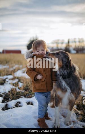 Hund leckt das Gesicht eines Kleinkindes draußen im Winter in Stockfoto