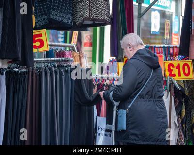 Käufer, die Preise und Waren auf dem Walthamstow Market, London, Großbritannien, dem längsten Outdoor-Markt in Europa, suchen. Händler und Inhaber von Verkaufsständen bedienen Kunden, wobei die Preise in Pfund angegeben werden Stockfoto