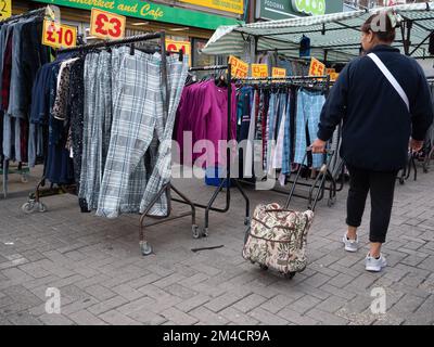 Käufer, die Preise und Waren auf dem Walthamstow Market, London, Großbritannien, dem längsten Outdoor-Markt in Europa, suchen. Händler und Inhaber von Verkaufsständen bedienen Kunden, wobei die Preise in Pfund angegeben werden Stockfoto