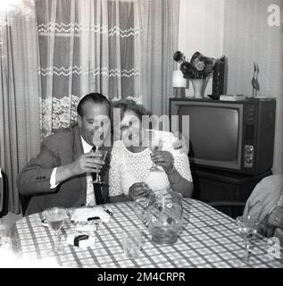 1960er Jahre, historisch, abends und ein reifer Mann und eine Dame sitzen an einem Tisch in einem Vorderzimmer, ein Glas Wein in der Hand, eine Zigarettenpackung auf dem Tisch, Netzvorhänge am Fenster und ein Fernsehgerät der Epoche in der Ecke, England, Großbritannien. Stockfoto