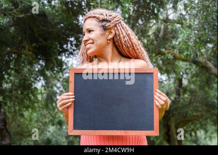 Eine lateinische Frau, die eine Tafel hält und nach links in einem Park schaut Stockfoto