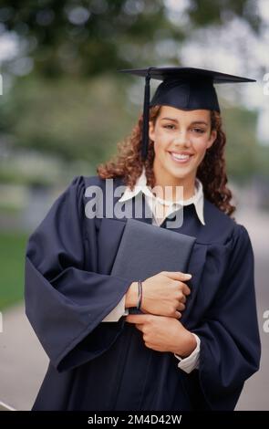 Ein afroamerikanisches Mädchen, das in Mütze und Kleid posiert und ihre Abschlussurkunde in der Hand hat, lächelt in die Kamera Stockfoto