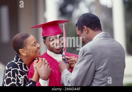 Afroamerikanische Mutter und Vater stehen mit ihrem Sohn, der gerade seinen Abschluss gemacht hat, und richten seine Krawatte. Alle lächeln und sind glücklich Stockfoto
